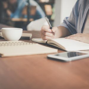 woman writing on a notebook beside teacup and tablet computer