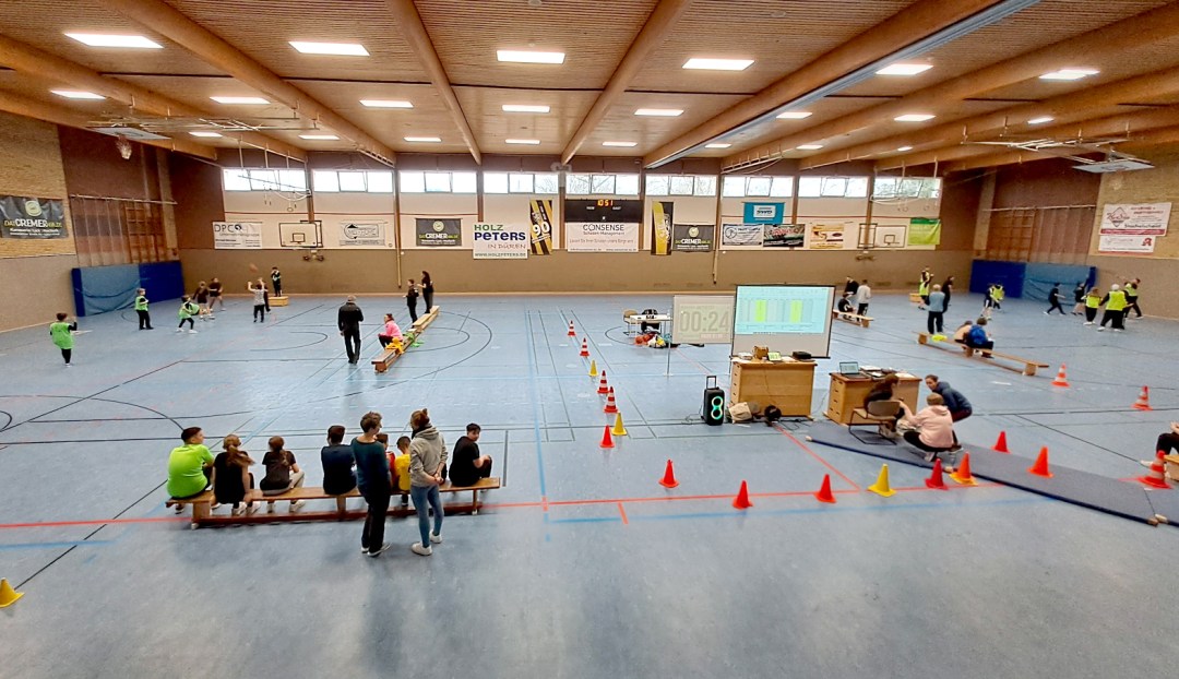 Sporthalle mit mehreren Personen, die an einem Basketballturnier teilnehmen, und Zuschauern auf einer Bank.