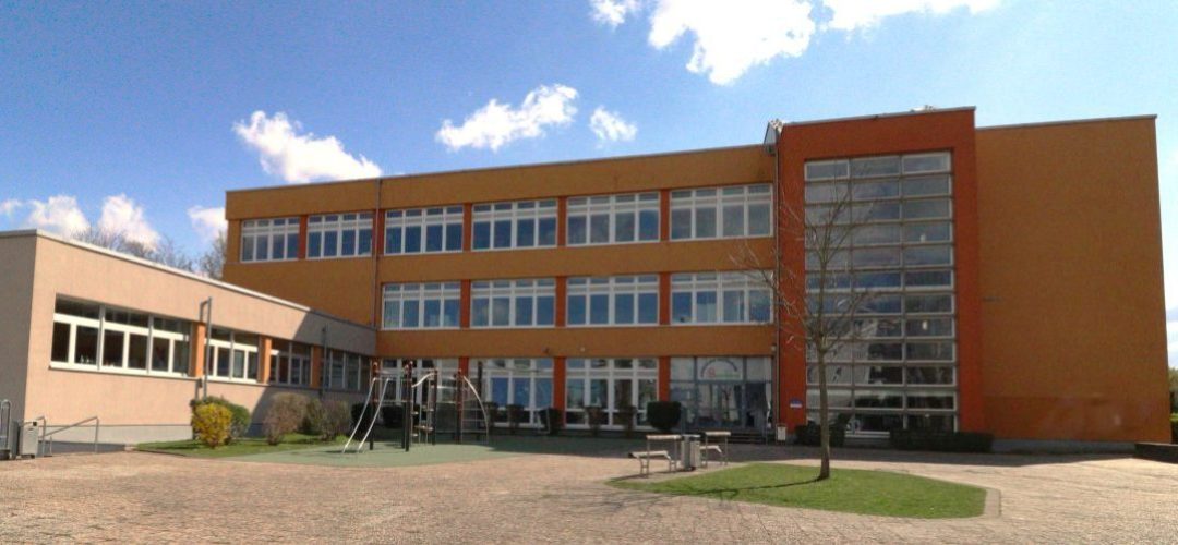 Schulgebäude mit orangefarbenen und grauen Fassaden und einem Spielplatz im Vordergrund.